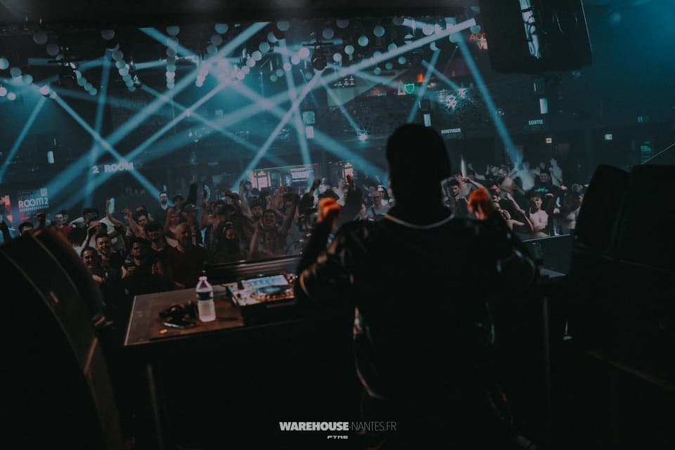 Vue depuis la régie à la Fête de la Hard Music : silhouette de l'artiste bras levés face à la foule, croisillons de beams blancs, Warehouse Nantes